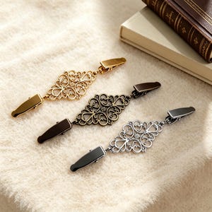 Puede incluir: Tres pinzas decorativas de metal para chal en oro, bronce y plata. Cada pinza presenta un diseño de filigrana ornamental y simétrico y un cierre rectangular. Las pinzas se muestran sobre una tela color crema.