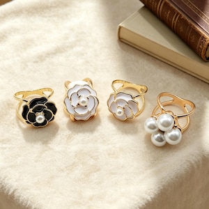 May include: Four gold-tone scarf rings with floral and pearl designs. One ring features a black flower with a pearl center, two have white flowers, and one has a cluster of pearls.