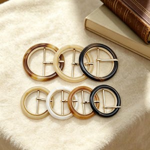 May include: A collection of seven round belt buckles in various colors, including brown, beige, white, and black. Each buckle features a gold-colored metal bar across the center. The buckles are arranged on a soft, cream-colored surface.