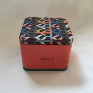 Może przedstawiać: Czerwono-czarne pudełko z nadrukiem "Fossil" na przodzie.