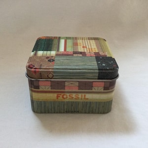 Op de afbeelding: Een vierkante, gepatenteerde blikken doos met het woord "FOSSIL" erop gedrukt. De doos is gemaakt van metaal en heeft een kleurrijk geometrisch ontwerp.