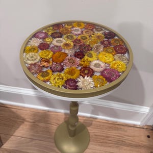 May include: A round side table with a clear resin top embedded with colorful dried flowers. The table has a gold-colored rim and a matching pedestal base. The table is approximately 24 inches tall.