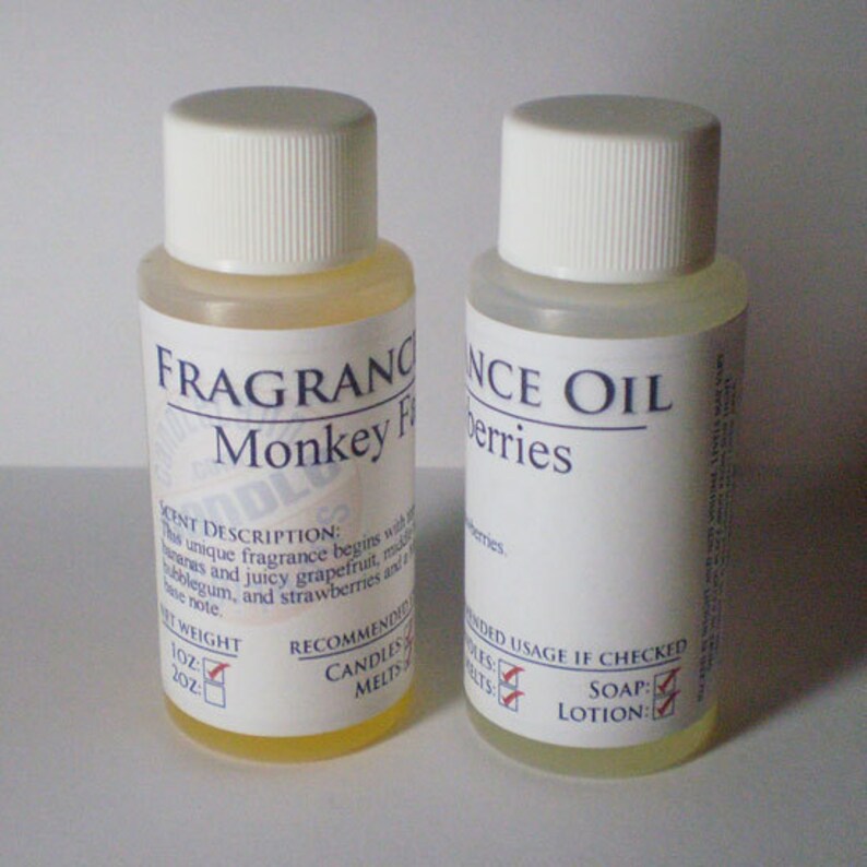 May include: Two small clear glass bottles with white labels. The bottle on the left is labeled "FRAGRANCE OIL Monkey Fruit" and the bottle on the right is labeled "FRAGRANCE OIL Berries". Both bottles have a scent description, recommended usage, and net weight listed on the label.