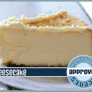 May include: A slice of cheesecake with a white frosting and a brown crust. The cheesecake is on a white plate with a blue and white sticker that says "approved" and "seamz".