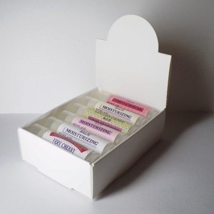 May include: A white cardboard display box filled with several lip balm tubes. The tubes have labels with the words "Moisturizing Balm" and various flavors like "Very Cherry" and "Cotton Candy". The box is open, showcasing the products.