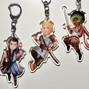 May include: Three acrylic keychains with fantasy character designs. Each keychain has a silver metal clasp. One character is an elf with long black hair and a blue dress. Another is a blonde man in armour. The third is a person of colour holding a sword.