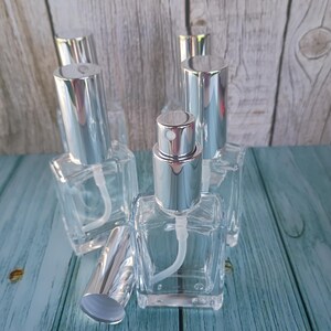 May include: Clear glass perfume bottles with silver-colored spray tops and caps. The bottles are square-shaped and have a white plastic tube for dispensing the liquid. The caps are cylindrical and sit on top of the bottles. The bottles are arranged on a blue wooden surface.