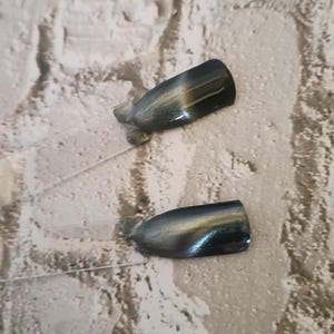 Puede incluir: Dos paletas de plástico transparente para exhibir esmalte de uñas con un acabado gris carbón metálico oscuro y un sutil efecto magnético de ojo de gato dorado brillante que crea una franja de luz diagonal en la superficie de cada uña.