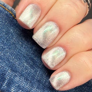 Puede incluir: Uñas pintadas con un esmalte plateado brillante y reflectante que muestra sutiles tonos holográficos verdes y rosados. Las uñas tienen bordes redondeados y se presentan sobre un fondo de tela de mezclilla azul texturizada.