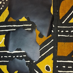 Può includere: Ritagli di pelle nera a forma di continente africano sono posizionati su un tessuto fantasia. Il tessuto presenta un design geometrico giallo, nero e bianco. I ritagli si sovrappongono leggermente.