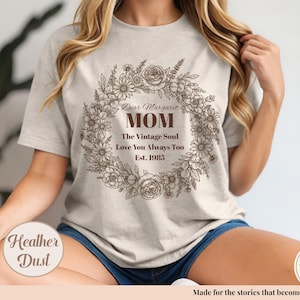 Pu&ograve; includere: Una maglietta color polvere di erica con un design a ghirlanda floreale e le parole "Dear Margaret MOM" e altro testo. La maglietta include anche il testo "The Vintage Soul Love You Always Too Est. 1985".
