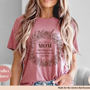Pu&ograve; includere: T-shirt color malva heather con un design a ghirlanda floreale. Il testo recita "Dear Margaret MOM The Vintage Soul Love You Always Too Est. 1983". La maglietta &egrave; in stile casual.