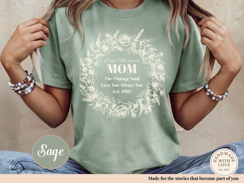 Pu&ograve; includere: T-shirt verde salvia con un design a ghirlanda floreale. Il testo sulla maglietta recita: "Dear Margaret MOM The Vintage Soul Love You Always Too Est. 1985". La maglietta &egrave; realizzata in un materiale morbido ed &egrave; comoda.