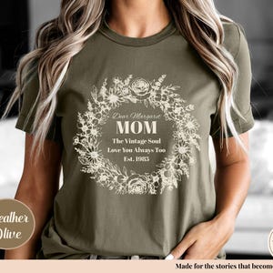 Pu&ograve; includere: T-shirt verde oliva con un design a ghirlanda floreale. Il testo recita "Dear Margaret MOM The Vintage Soul Love You Always Too Est. 1985". Un'etichetta rotonda dice "Heather Olive".