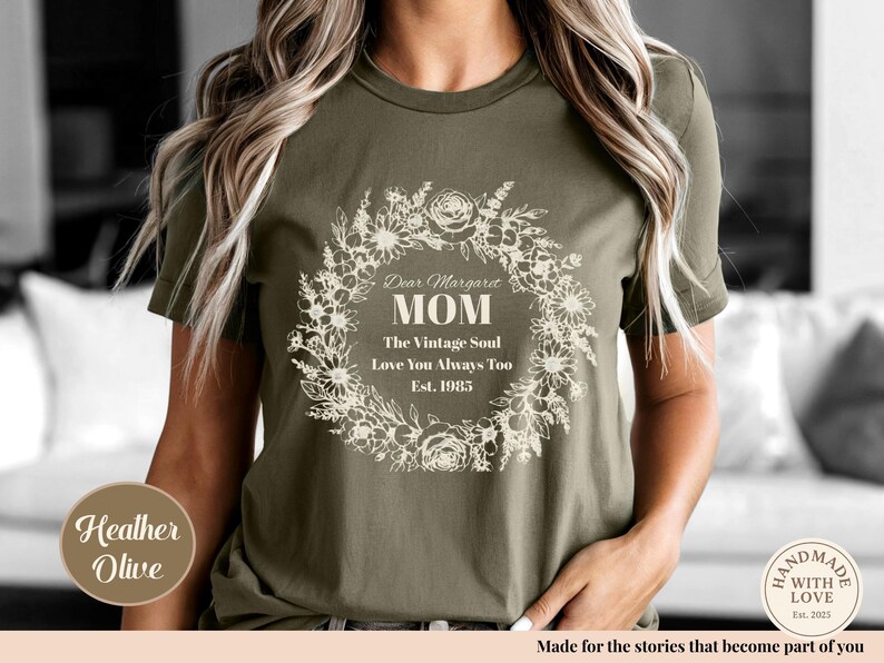 Pu&ograve; includere: T-shirt verde oliva con un design a ghirlanda floreale. Il testo recita "Dear Margaret MOM The Vintage Soul Love You Always Too Est. 1985". Un'etichetta rotonda dice "Heather Olive".