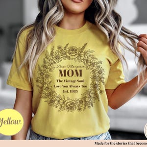 Pu&ograve; includere: T-shirt giallo senape con un design a ghirlanda floreale. Il testo all'interno della ghirlanda recita: "Dear Margaret Mom, The Vintage Soul, Love You Always Too, Est. 1985." Un cerchio giallo con la parola "Yellow" &egrave; in basso a sinistra.
