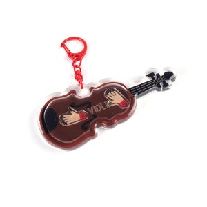 Pode incluir: Um chaveiro em forma de violino marrom com fecho vermelho. O chaveiro apresenta um design de violino com a palavra "VIOLI" impressa. O design do violino inclui ilustrações de mãos com unhas pintadas de vermelho.