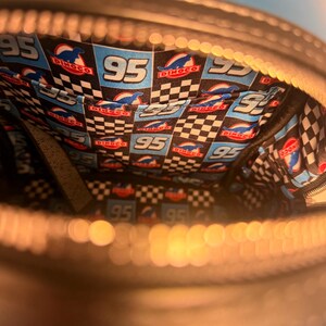 May include: Black zippered bag with a vibrant interior featuring a checkered pattern and the number "95" in white. The interior also displays the "Dinoco" logo and a blue race car design. The bag has multiple pockets.