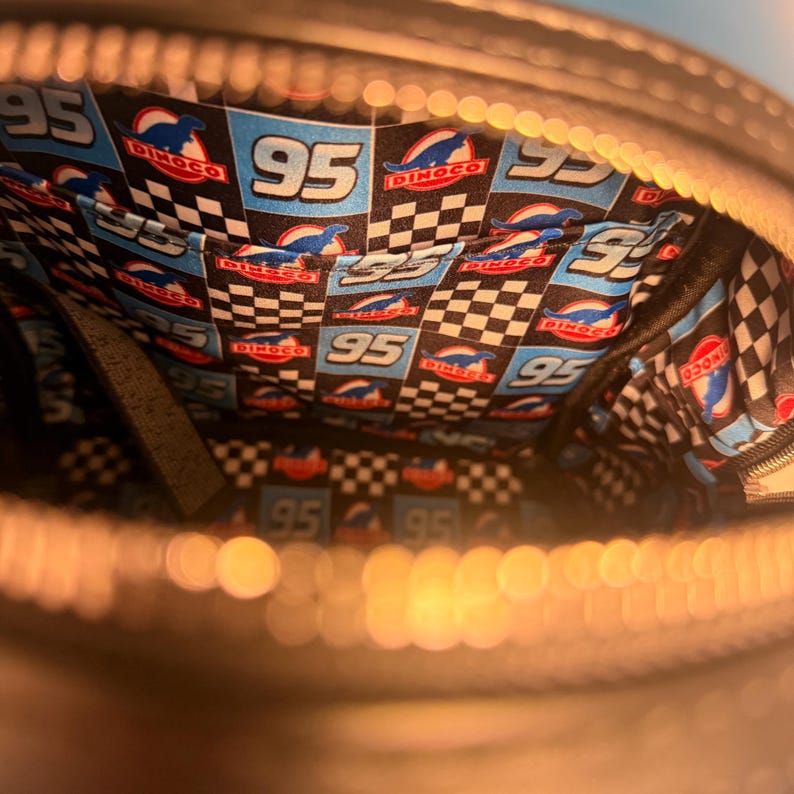 May include: Black zippered bag with a vibrant interior featuring a checkered pattern and the number "95" in white. The interior also displays the "Dinoco" logo and a blue race car design. The bag has multiple pockets.