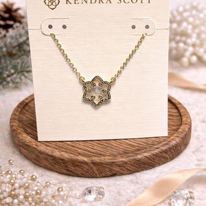 Könnte beinhalten: Eine goldfarbene Halskette mit einem Schneeflocken-Anhänger, präsentiert auf einer cremefarbenen Karte mit dem Logo "Kendra Scott". Die Halskette wird auf einem Holztablett mit dekorativen Elementen im Hintergrund präsentiert.