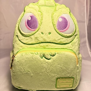 Könnte beinhalten: Ein grüner Plüschrucksack in Form eines Cartoon-Gesichts mit großen, gestickten Augen mit rosa und lila Details. Der Rucksack hat eine Vordertasche mit Reißverschluss und einen goldfarbenen Reißverschluss. Ein kleines rechteckiges Etikett mit "Disney Parks" und "loungefly" ist sichtbar.
