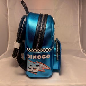 May include: A blue backpack with a shiny finish, featuring black zippers and a checkered pattern. The backpack has the word "DINOCO" and the number "95" printed on it, with a racing theme.