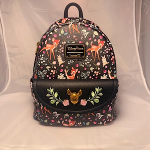 May include: Black mini backpack featuring a Bambi design with deer and floral patterns. The backpack has a front flap with a gold deer head emblem and the text "Disney Parks Loungefly". The bag has a top handle and adjustable straps.