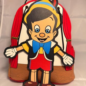 May include: Red backpack with a large, colorful, cartoon image of Pinocchio on the front. The backpack has a drawstring closure and a wooden stage design.