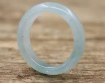 May include: A light blue jade ring. The ring is a solid band with a smooth, polished surface. The color is a pale, translucent blue-green, with subtle variations in tone. The ring is set against a wooden background.