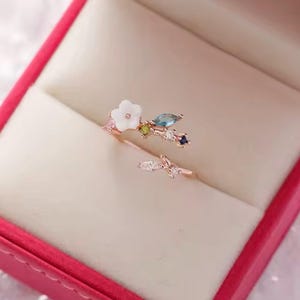 Pode incluir: Um anel de ouro rosa com um design floral, incluindo uma flor branca e pedras preciosas coloridas. O anel tem uma delicada estrutura em forma de ramo com haste dividida, apresentado numa caixa de joias rosa.
