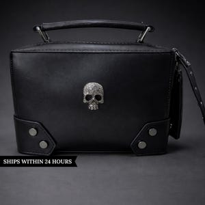 May include: Black rectangular handbag with a silver skull emblem. The bag has a top handle, corner studs, and a detachable strap. The bag is made of a smooth, matte material.