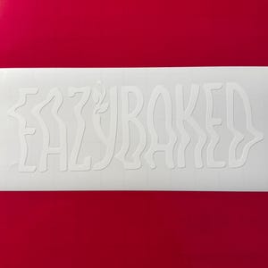 Puede incluir: Adhesivo de vinilo blanco con la palabra "EAZYBAKED" en una fuente ondulada y estilizada. La pegatina es rectangular y está colocada sobre un fondo rojo liso. Las letras tienen un diseño único y fluido.