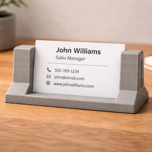 May include: A grey business card holder with a card displaying the name "John Williams" and contact information. The holder is rectangular with a slot to hold the card upright. The background is a wooden surface.