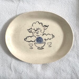 May include: Oval ceramic platter with a hand-painted sheep design in black and blue. The sheep is centered, with two wine glasses on either side. The platter has a speckled, off-white background.