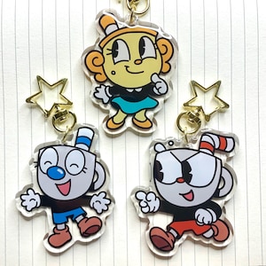 May include: Three clear acrylic keychains featuring cartoon characters with cup-shaped heads. One character is yellow with orange hair, one is white with a blue nose and shorts, and one is white with a red nose and shorts, all with star-shaped clasps.