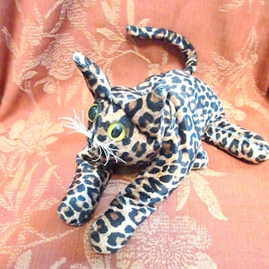 May include: A plush leopard cat toy with a spotted pattern in brown and black. The cat has large, yellow-eyed, and white whiskers. The toy is lying down with its legs extended and tail curved.