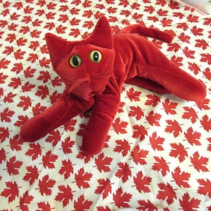 May include: A plush, red cat toy with large, round yellow eyes and a long, slender body. The cat is lying on a white fabric patterned with red maple leaves. The toy has a soft, velvety texture.