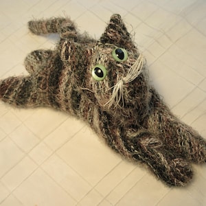 May include: A plush cat toy crafted from textured, variegated brown, grey, and black yarn. The figure features large, round, light green eyes with black pupils and long, thin, white thread whiskers extending from the snout area.