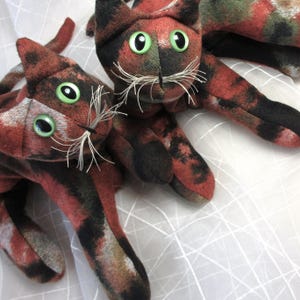 Peut inclure: Deux jouets pour chats en peluche faits à la main. Les chats sont faits d'un tissu à motifs dans les tons de rouge, noir et vert. Ils ont de grands yeux verts et des moustaches blanches. Les chats sont allongés sur une surface blanche.
