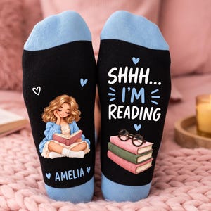 May include: A pair of black socks with light blue cuffs. The left sock features a cartoon girl with wavy hair reading a book and the name AMELIA. The right sock displays the text SHHH... I'M READING above a stack of three books and glasses.