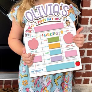 May include: A wooden sign with the text "Olivia's First Day Of" in purple and white. The sign has sections for favorites, including food, color, and animal. It also includes sections for height in inches and what the child wants to be when they grow up.