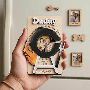Peut inclure: Un cadeau de fête des pères en bois en forme de barbecue, tenu dans une main. Le cadeau contient une photo et les mots "Daddy", "Happy Father's Day" et "Love, Anna". D'autres aimants en bois sont visibles.