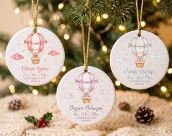 Op de afbeelding: Drie ronde witte keramische ornamenten hangend aan gouden koordjes. Elk toont een teddybeer in een heteluchtballon, wolken en sterren. Ze zijn gepersonaliseerd met namen, geboortegewicht, lengte en geboortedatum.