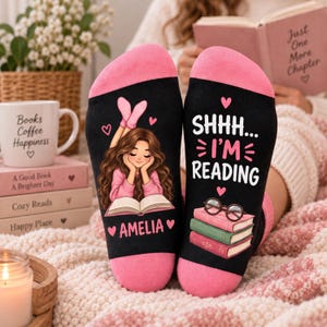 Op de afbeelding: Een paar zwarte sokken met roze hielen en tenen. Eén sok toont een illustratie van een persoon met lang bruin haar die een boek leest, met de naam Amelia eronder. De andere sok toont de tekst SHHH... I'M READING en een stapel boeken.