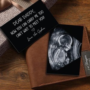 Puede incluir: Una tarjeta metálica negra con el texto "DEAR DADDY, NOW YOU CAN CARRY ME TOO CAN'T WAIT TO MEET YOU! Love, the Sophia" y una imagen de ultrasonido. La tarjeta está colocada sobre una cartera de cuero marrón.