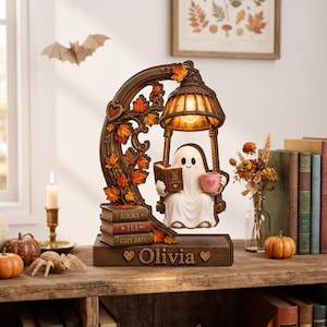 Puede incluir: Una lámpara decorativa con un fantasma blanco sentado en un columpio, leyendo un libro y sosteniendo una taza rosa. La base es marrón con el nombre Olivia grabado, y el marco está adornado con hojas de otoño naranjas y libros apilados.