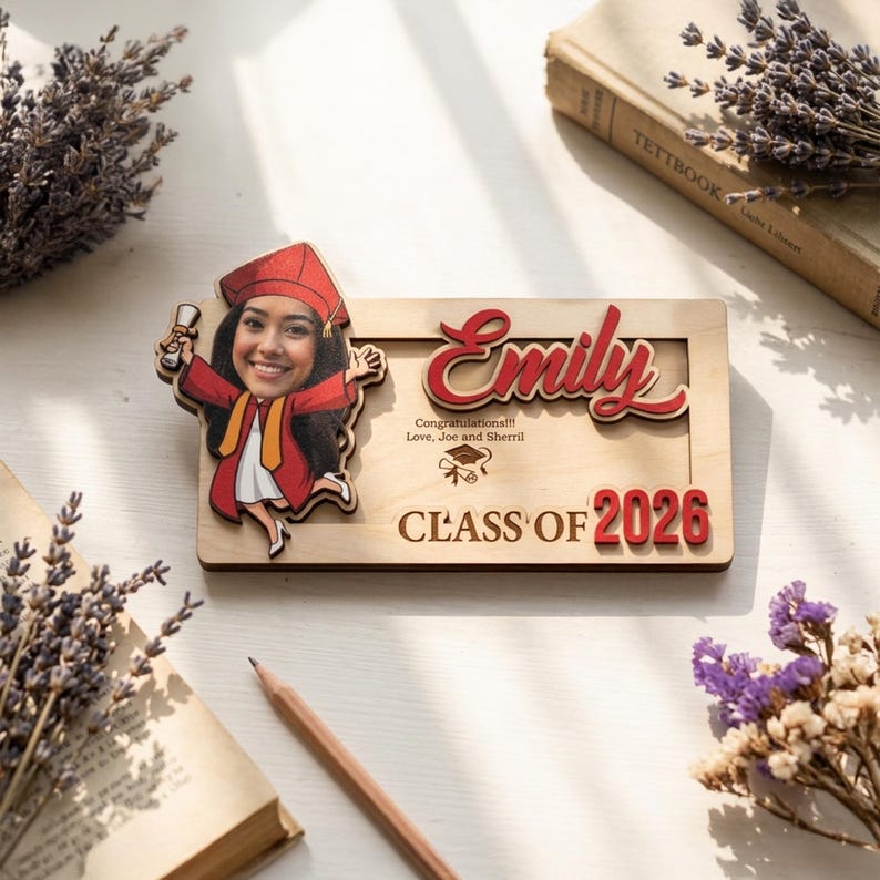 May include: A personalized graduation plaque featuring a photo of a graduate in a red cap and gown. The plaque includes the name "Emily" in red script, the text "Class of 2026," and congratulatory messages. The wooden plaque is rectangular.