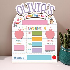 May include: A personalized wooden sign for the first day of school, featuring the name "Olivia's" in purple script. The sign includes sections for "Favorites" and other details, with a pink apple graphic and a red apple at the bottom.