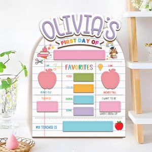 May include: A personalized wooden sign for the first day of school, featuring the name "Olivia's" in purple letters. The sign includes sections for "Favorites," "Years Old," and "Inches Tall," with a pink apple graphic and a red apple graphic.