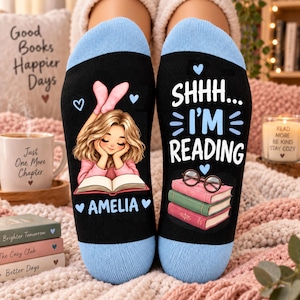 May include: A pair of black socks with light blue cuffs. The left sock features a cartoon girl with blonde hair reading a book, the name AMELIA, and small blue hearts. The right sock displays the text SHHH... I'M READING, blue hearts, and a stack of three books topped with round glasses.